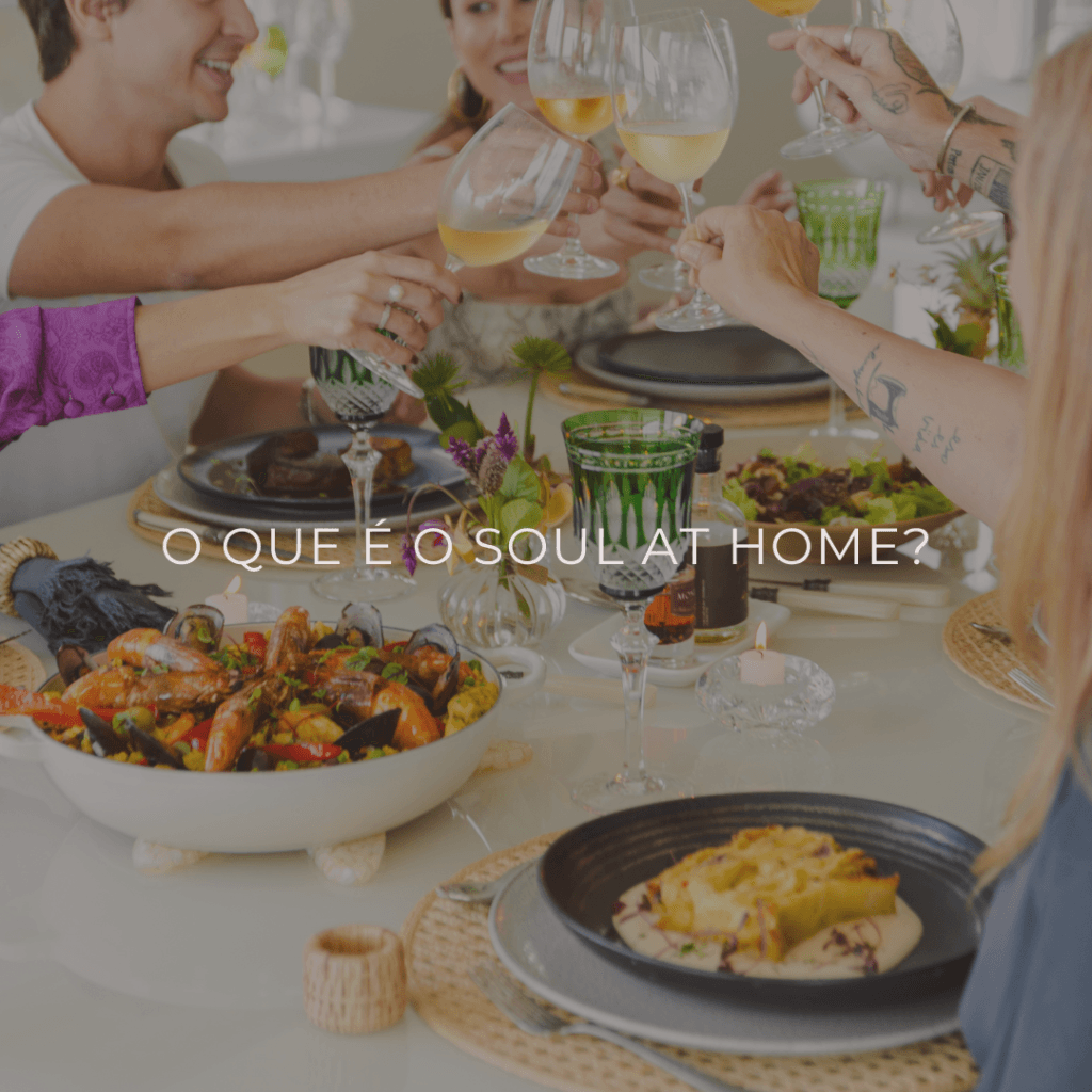 Mesa elegantemente posta com pratos e uma paella com frutos do mar refinados e taças de vinho, em um ambiente sofisticado. Ao fundo, um grupo de amigos brinda e conversa, criando uma atmosfera descontraída e intimista. A composição da imagem transmite exclusividade e celebração, refletindo o conceito de alta gastronomia do Soul At Home.